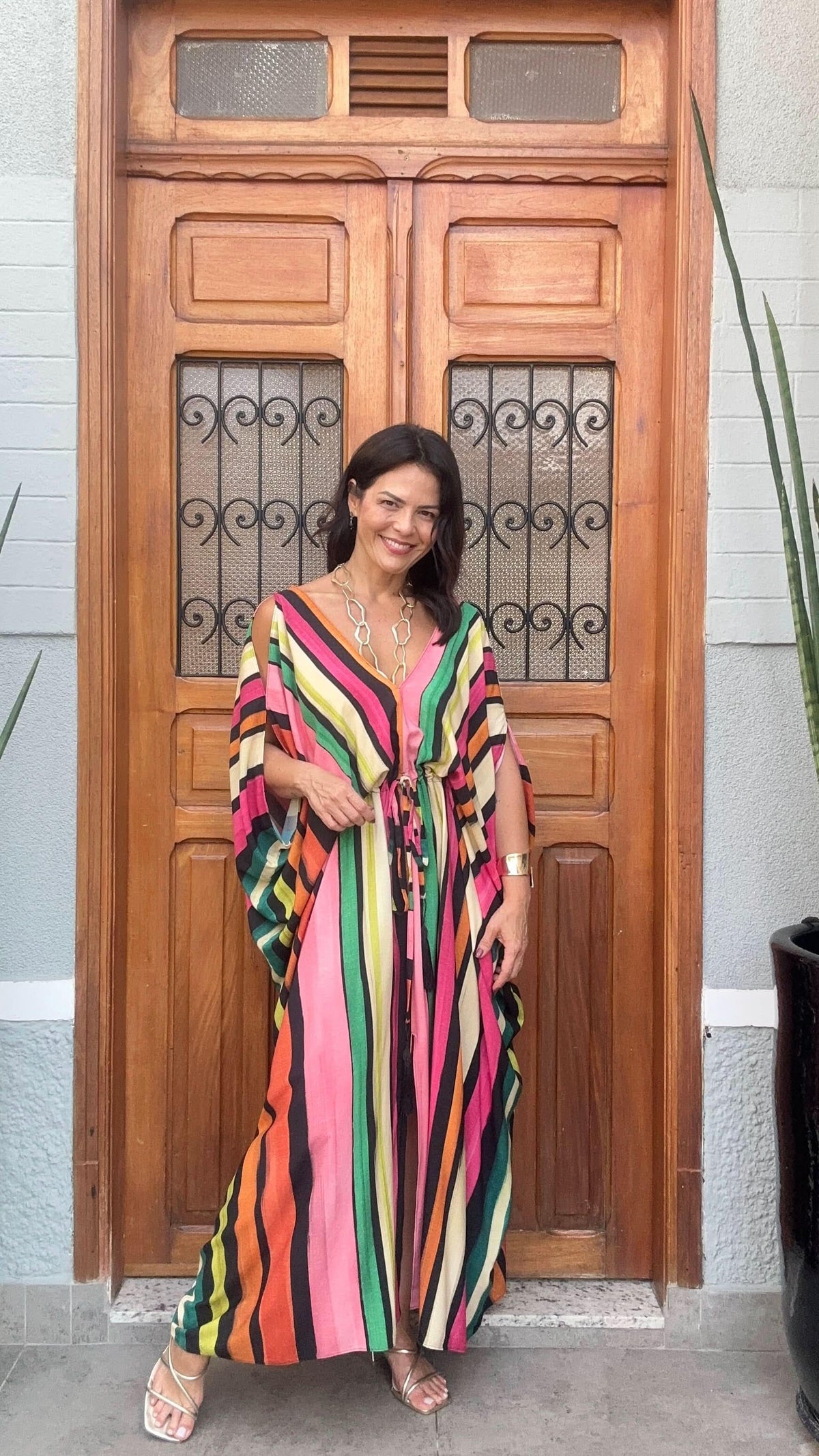 Mulher sorrindo usa kaftan longo listrado colorido em frente a porta de madeira decorada.
