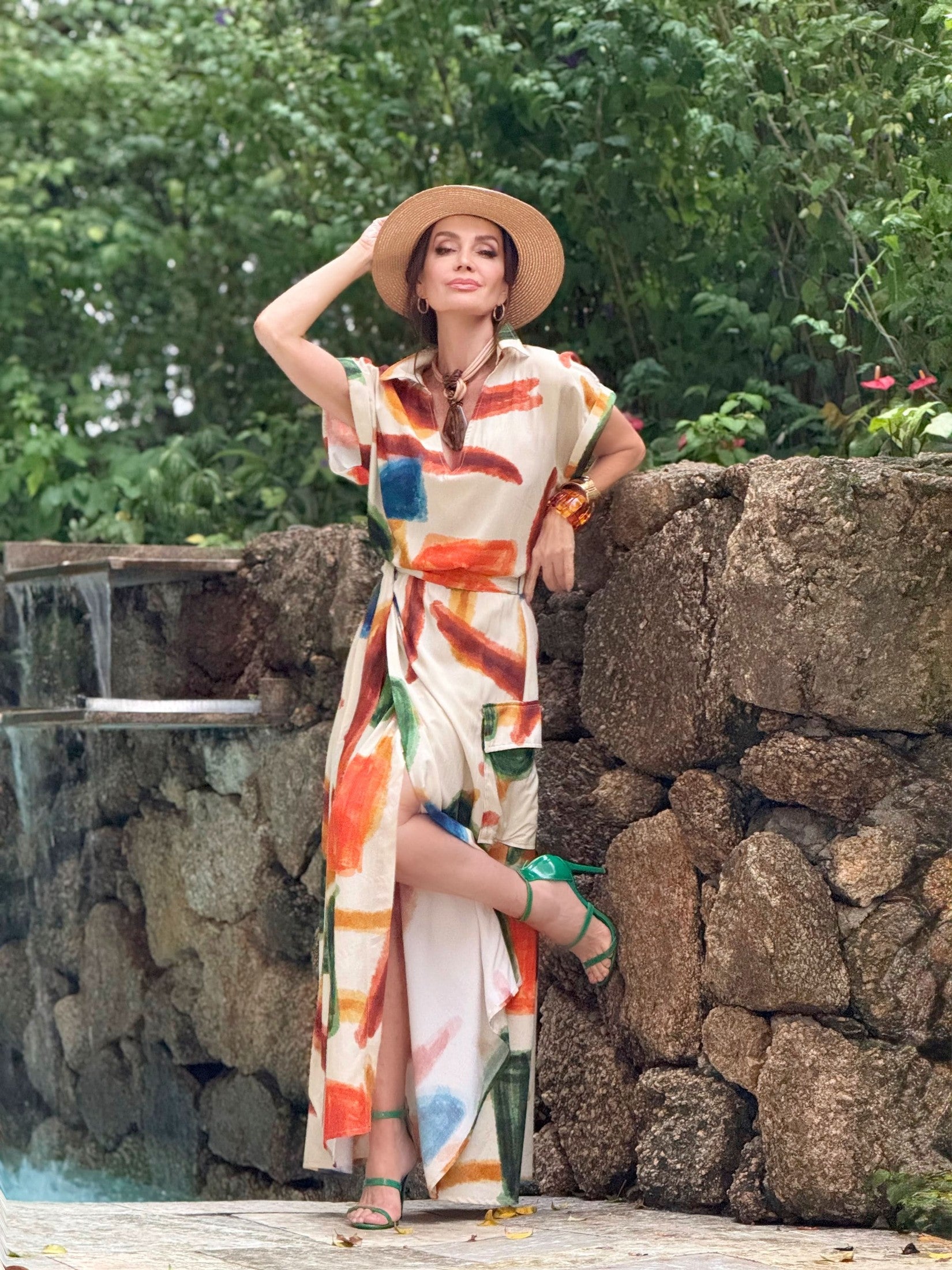 Mulher usando conjunto de saia com estampa abstrata colorida, chapéu e sandálias verdes, posando ao ar livre junto a um muro de pedras.