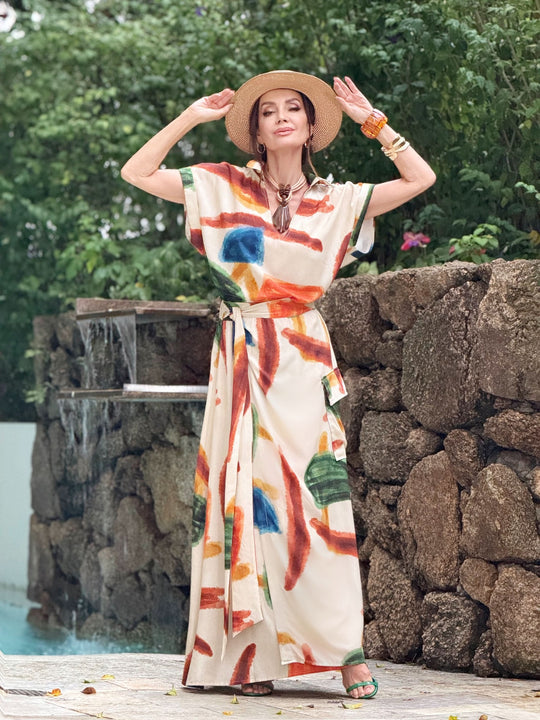 Mulher com saia longa estampada colorida e chapéu, em cenário ao ar livre com pedras e vegetação.