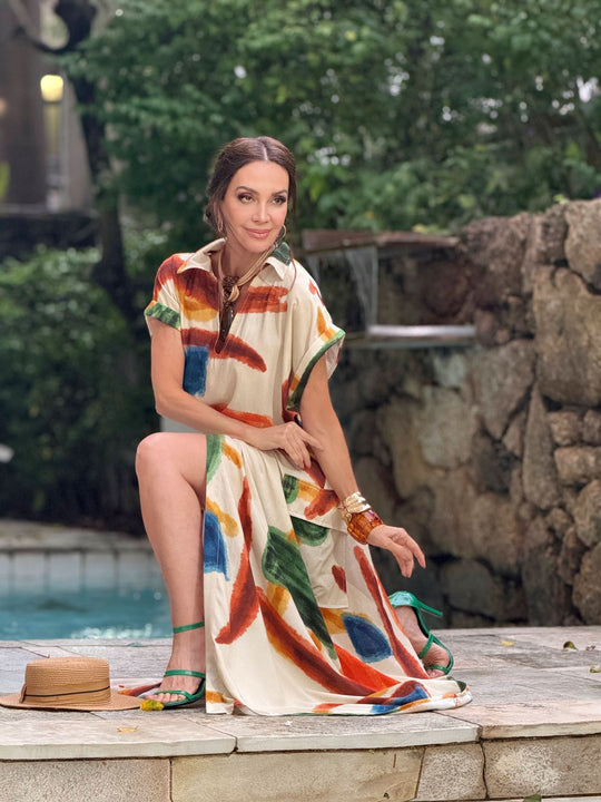 Mulher usando vestido longo estampado com traços coloridos, sentada ao ar livre perto de piscina e pedras.