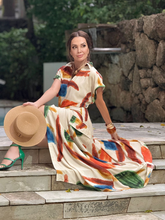 Mulher usando conjunto de saia longa com estampas coloridas, sentada em escada ao ar livre, segurando chapéu.