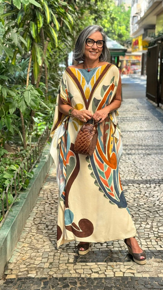 Mulher usando kaftan com estampa colorida, bolsa marrom e sandálias, em calçada urbana arborizada.