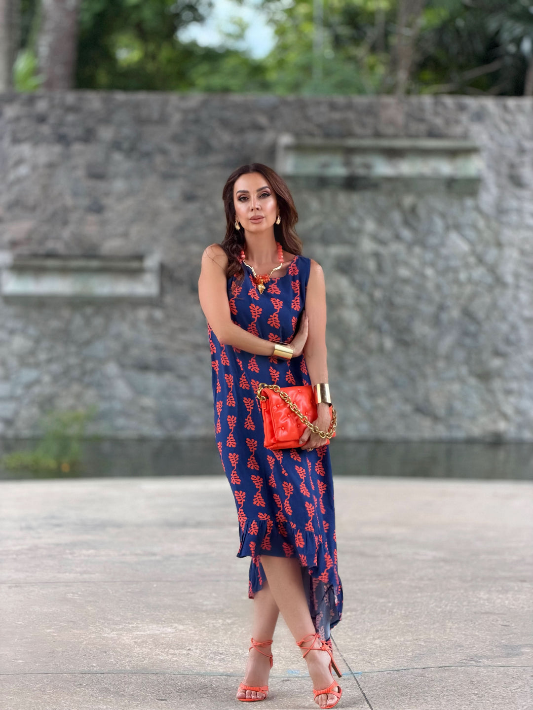 Mulher usando vestido azul estampado com folhas vermelhas, bolsa e sandálias laranja.