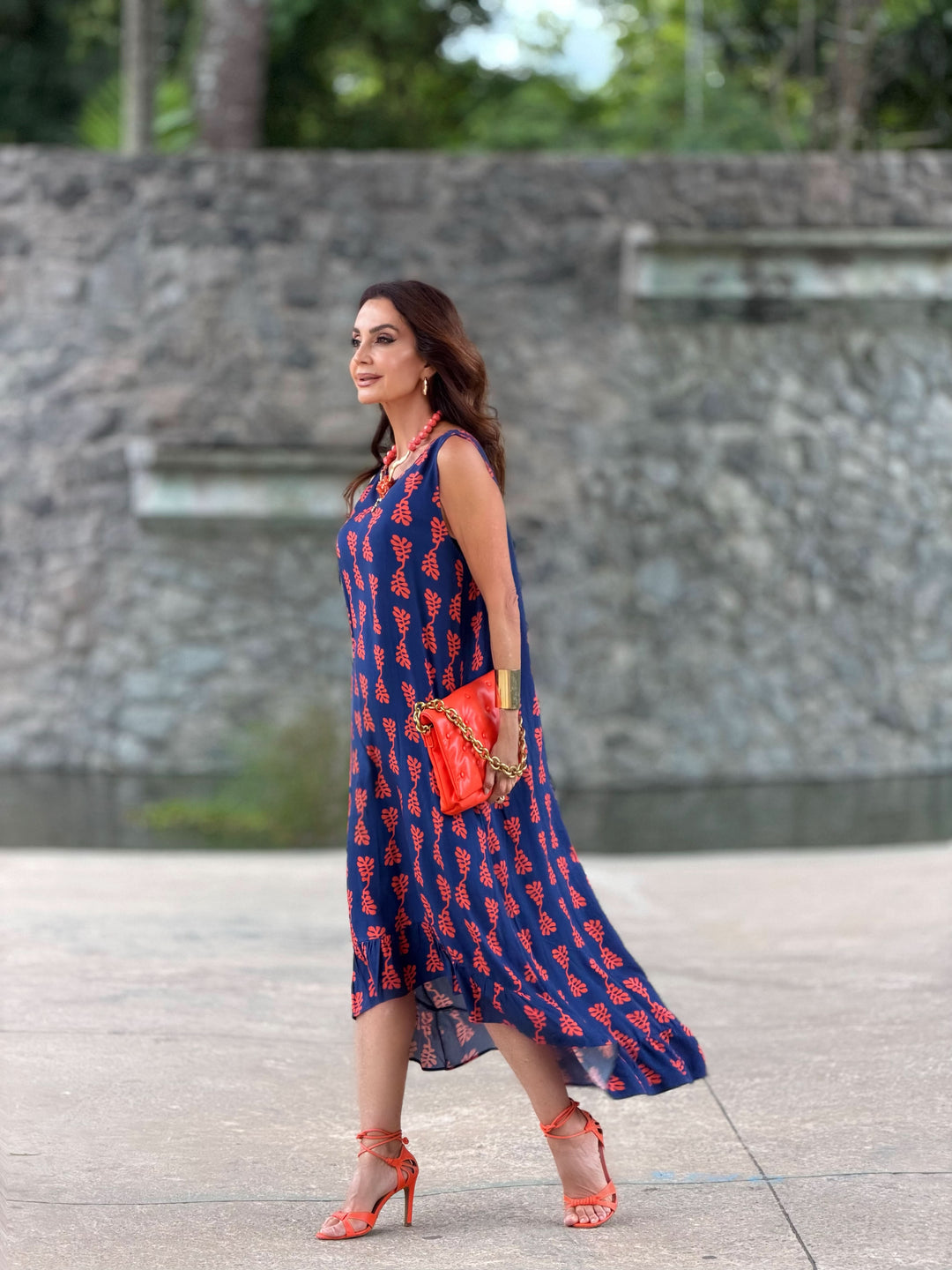 Mulher usando vestido longo azul com estampa vermelha, sandálias e bolsa laranja.