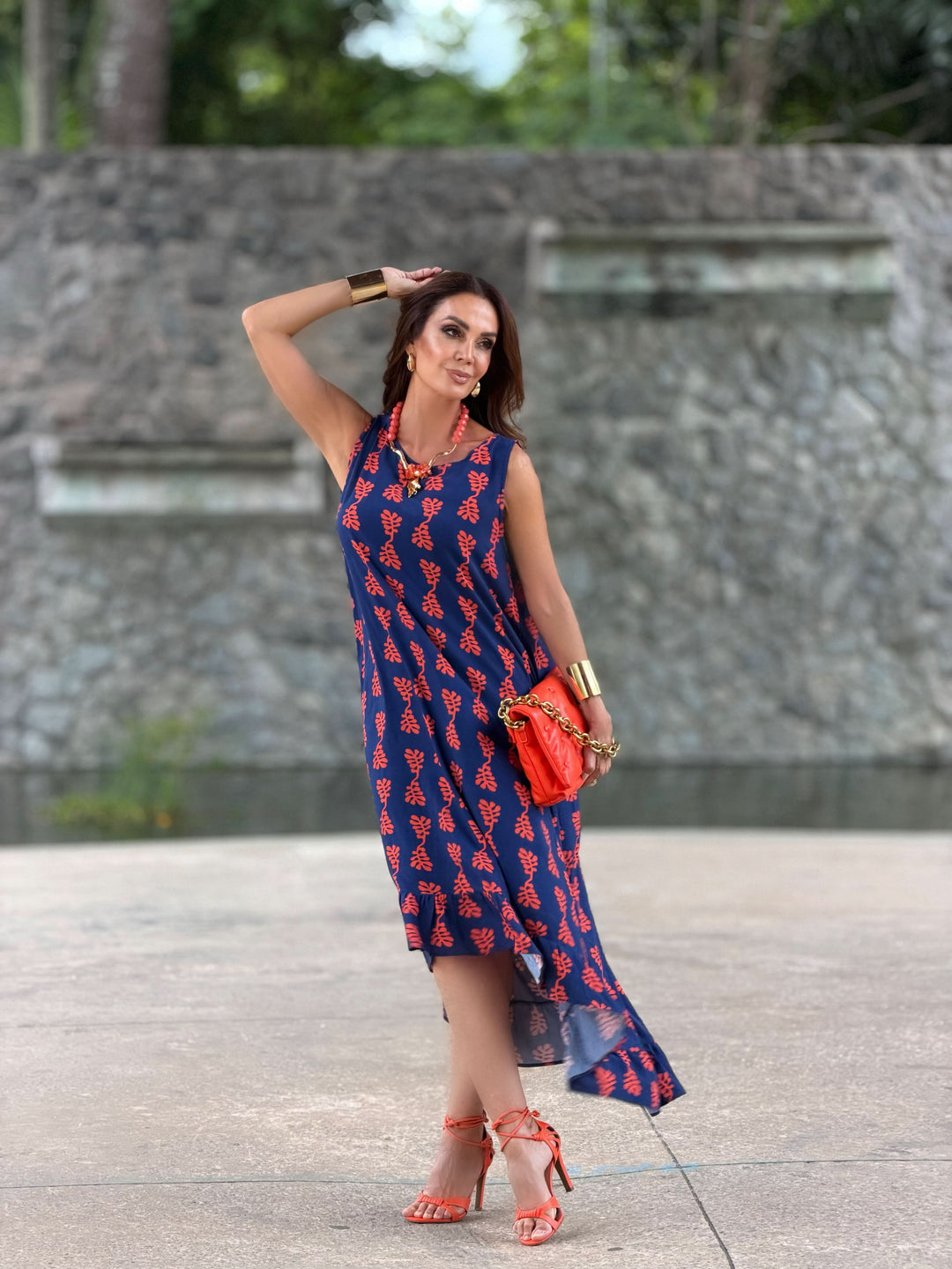 Mulher usando vestido midi azul estampado, sandálias e bolsa laranja, ao ar livre.