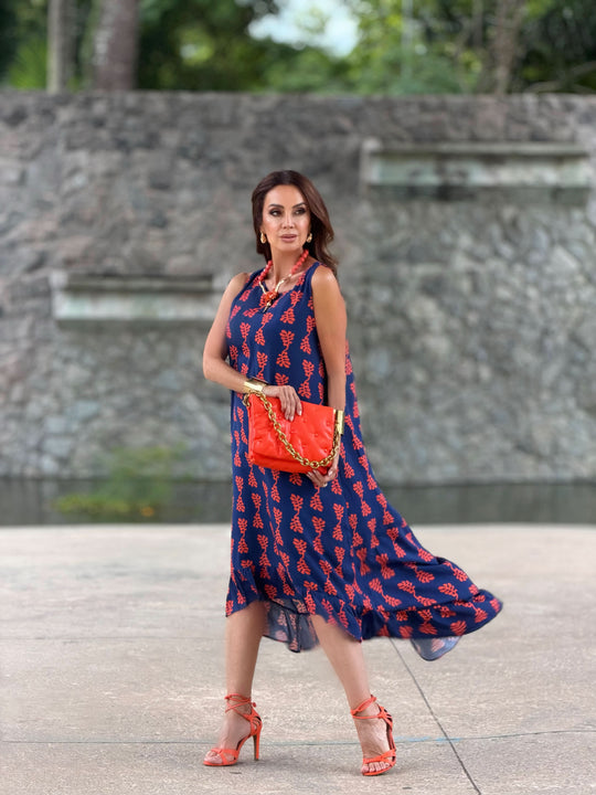 Mulher usando vestido midi azul estampado com folhas vermelhas, bolsa e sandália laranja, ao ar livre.