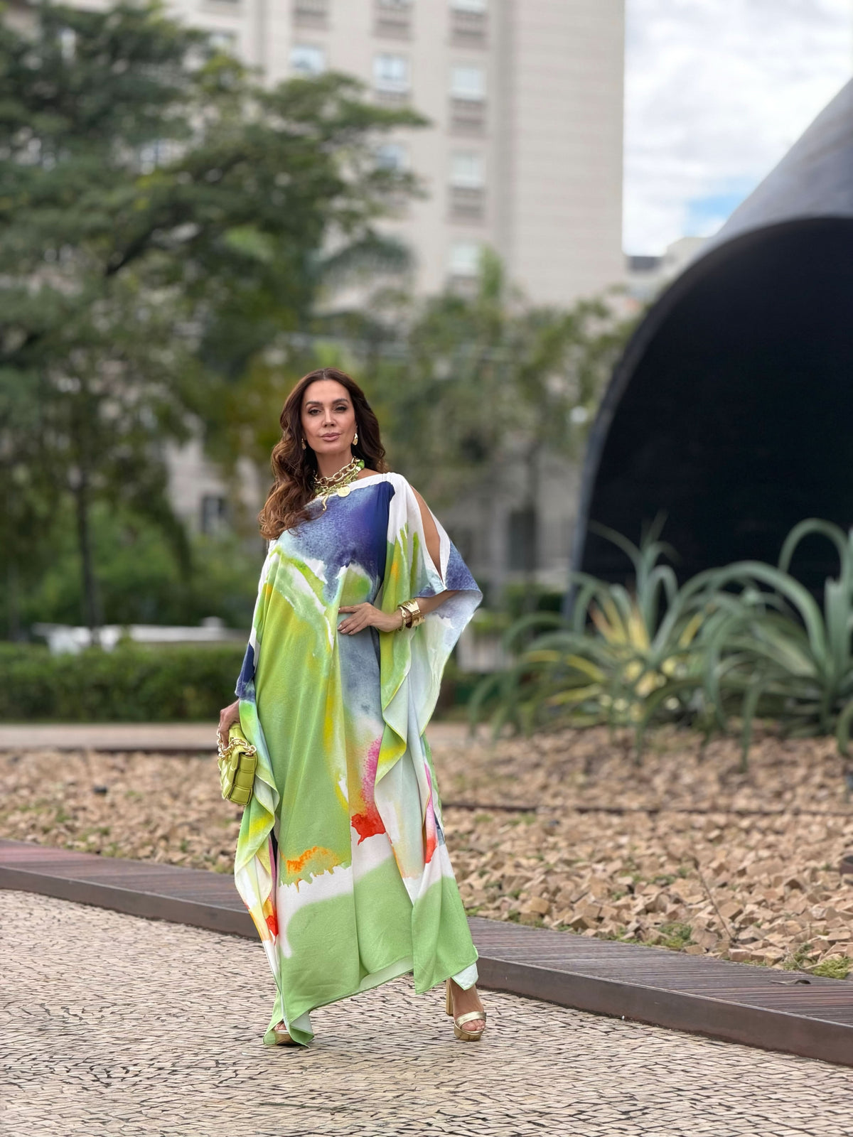 Mulher usando vestido kaftan longo estampado em tons de verde, azul e laranja, ao ar livre.