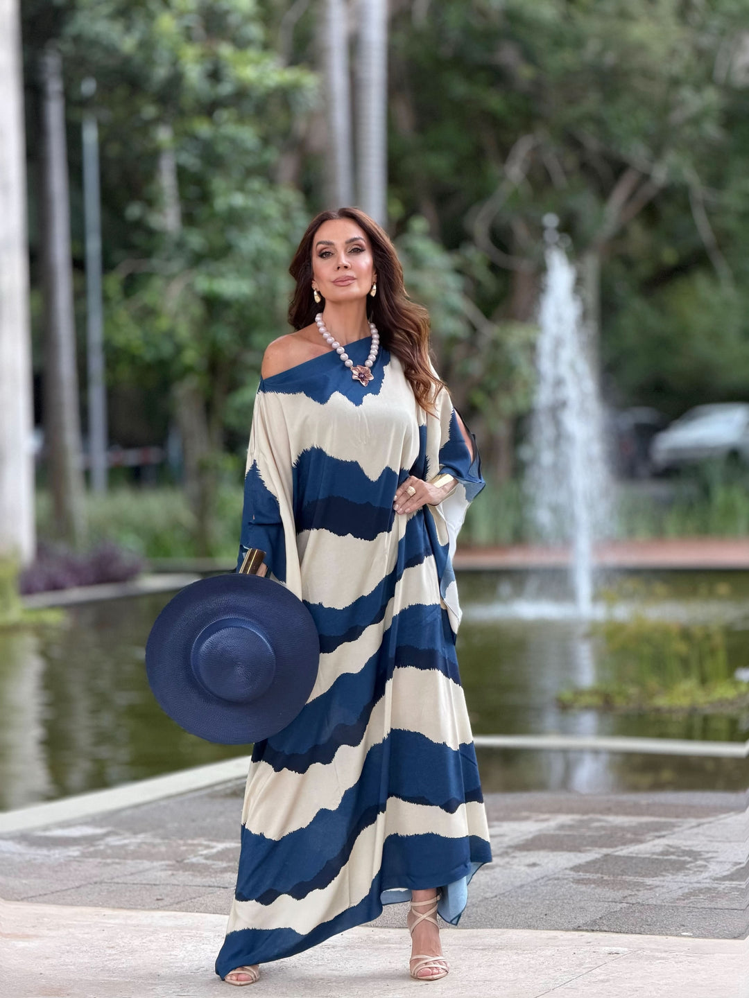 Mulher usando vestido kaftan longo azul e bege, segurando chapéu, ao ar livre junto a fonte.