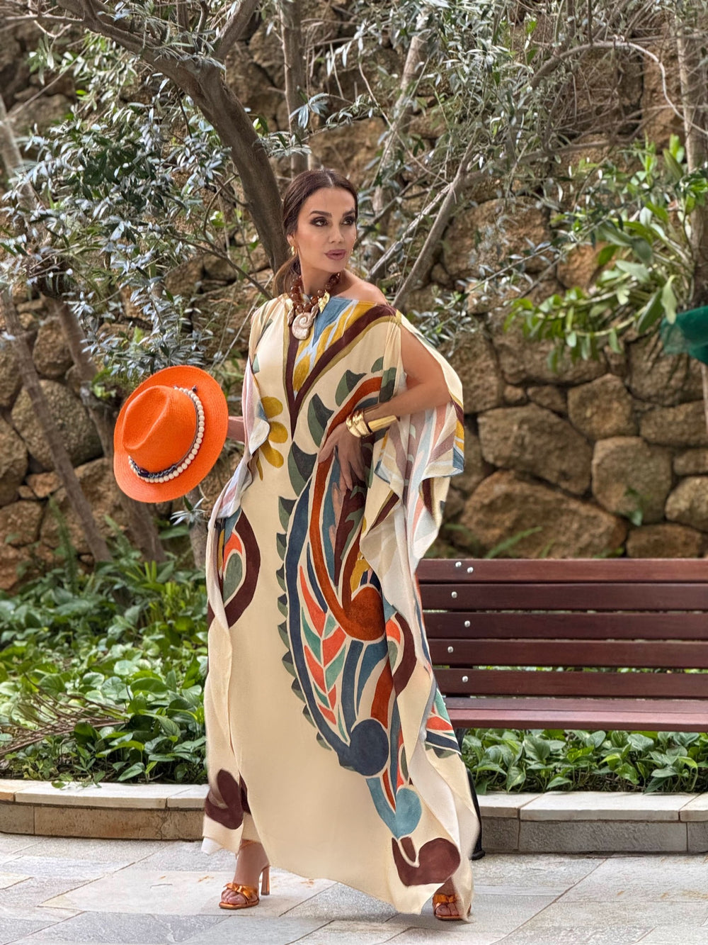 Mulher usando vestido kaftan estampado longo em seda, segurando chapéu laranja, em jardim