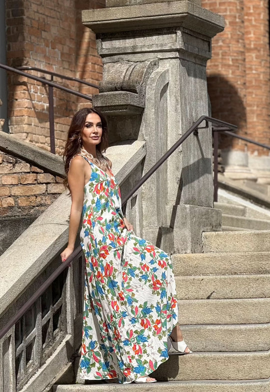 Mulher com vestido longo de alça, estampa floral colorida, em escadaria de pedra.