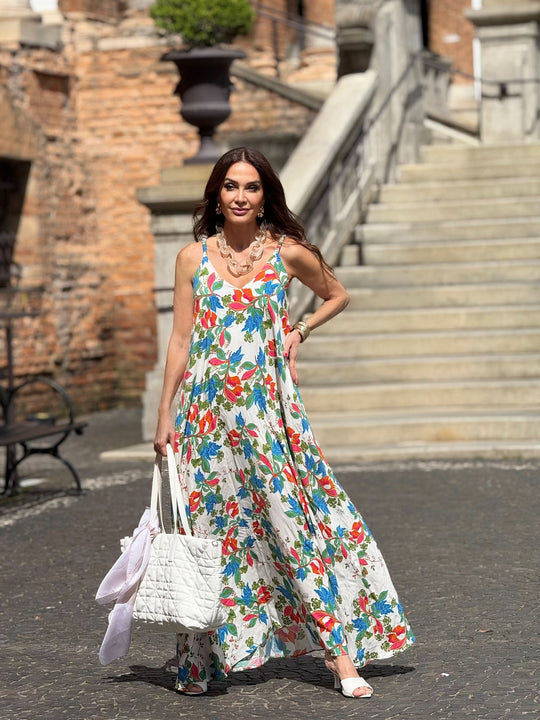 Mulher usando vestido longo de alça com estampa floral colorida, bolsa branca, ao ar livre.