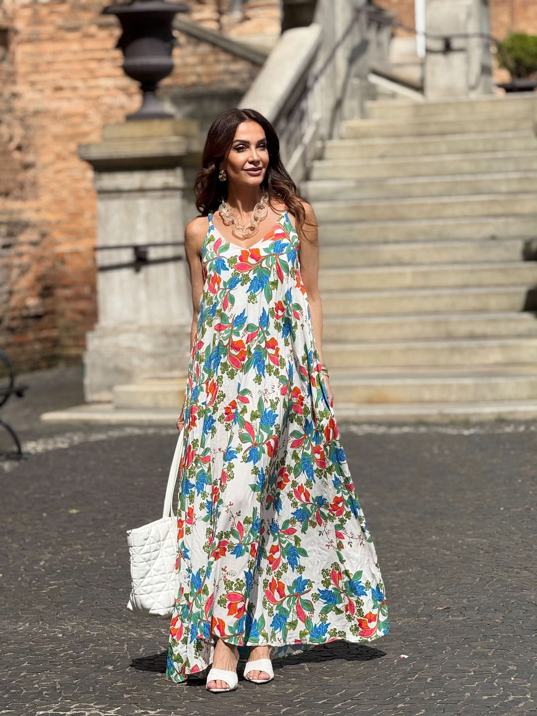 Mulher com vestido longo de alça, estampado floral colorido, bolsa branca e sandália, em ambiente externo com escadaria de pedra ao fundo.