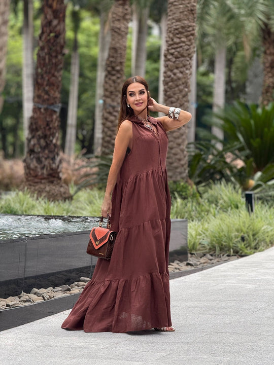 Mulher usando vestido longo de linho marrom, bolsa pequena e acessórios, ao ar livre com palmeiras.