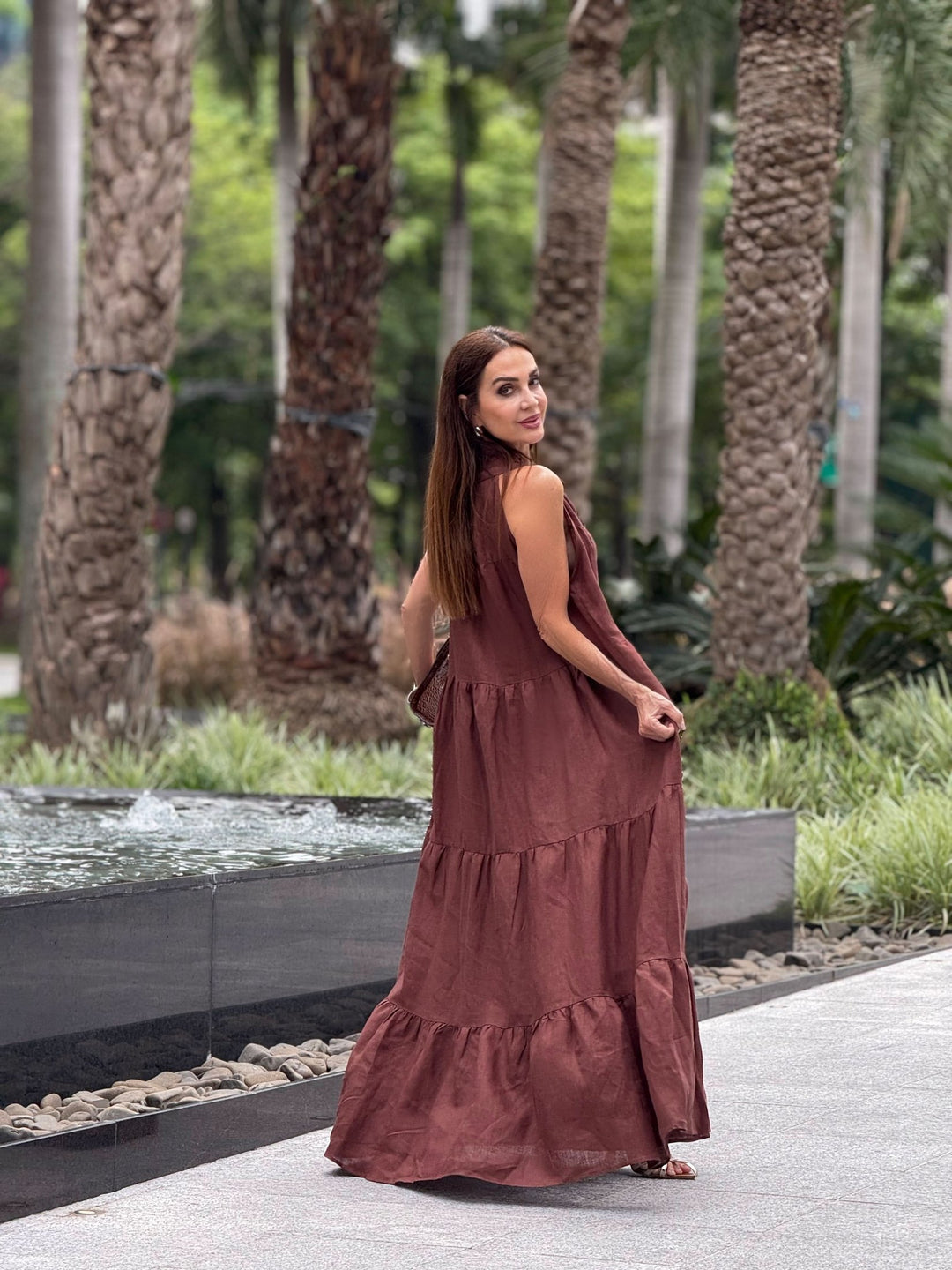 Mulher usando vestido longo de linho marrom ao ar livre, próximo a fonte e palmeiras.