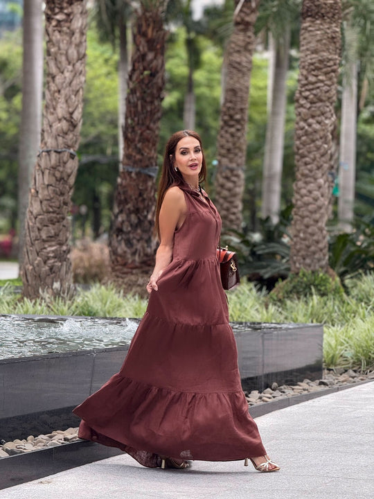 Mulher usando vestido longo de linho marrom, sandália de salto, em ambiente ao ar livre com palmeiras.