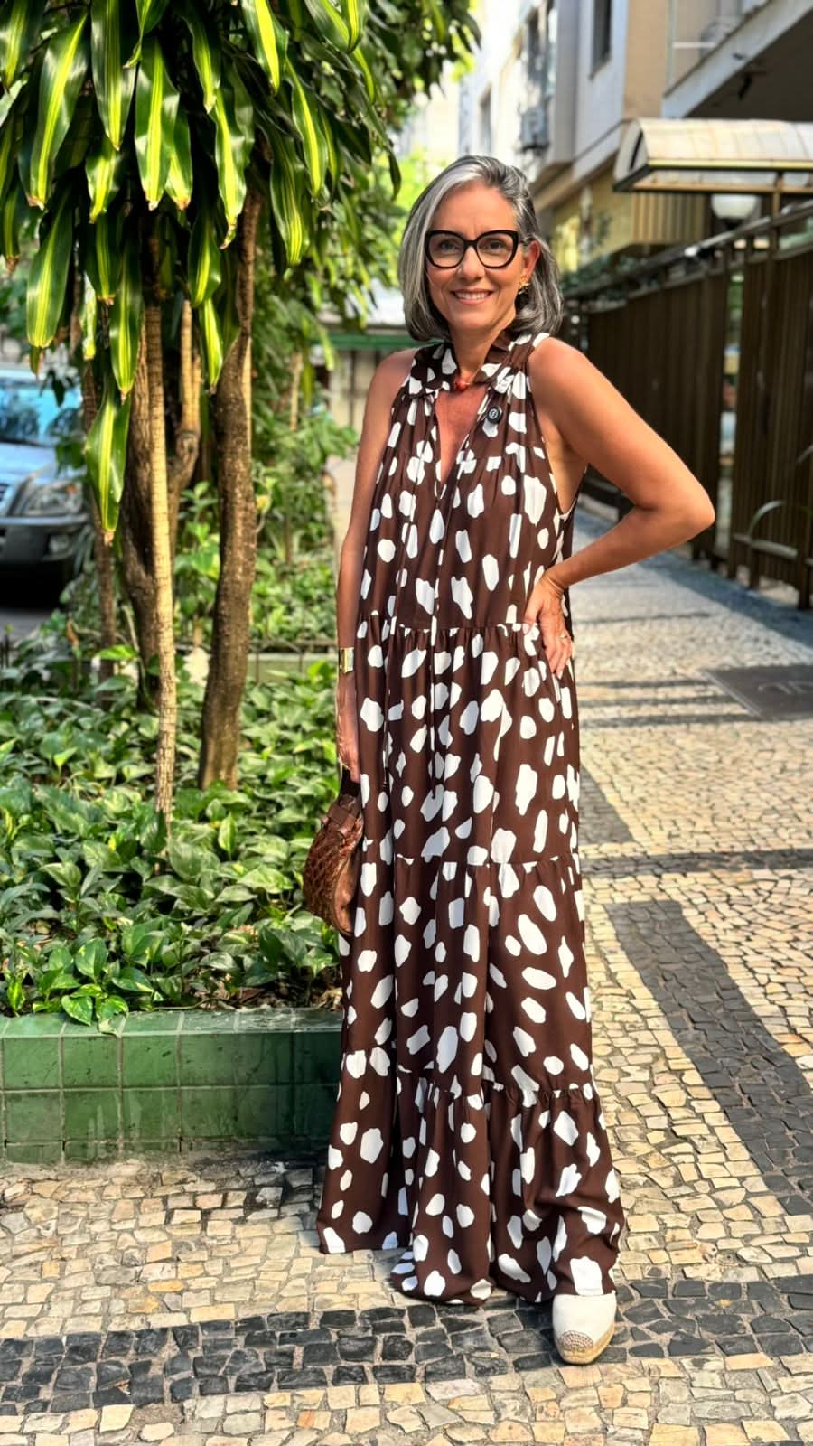 Mulher usando vestido longo marrom com estampa branca, em calçada ao ar livre.